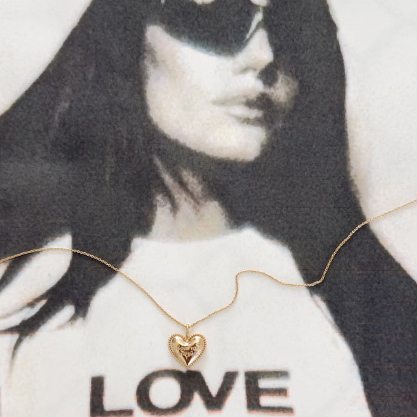 A delicate gold heart pendant necklace on a fine chain, displayed across a black-and-white fashion print featuring a woman in sunglasses and the word 'LOVE' on a white shirt.