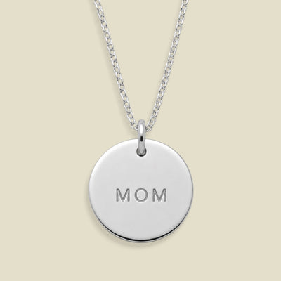 A silver round disc pendant necklace engraved with the word 'MOM', suspended from a delicate silver chain against a solid light beige background.