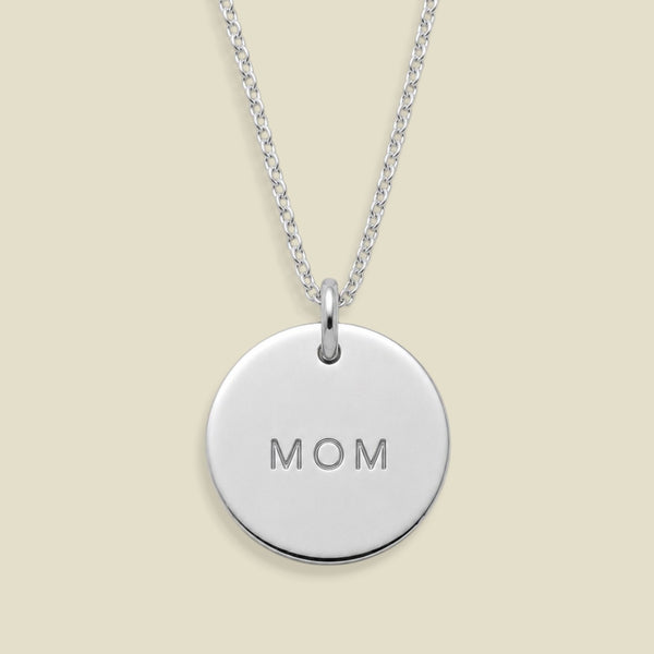 A silver round disc pendant necklace engraved with the word 'MOM', suspended from a delicate silver chain against a solid light beige background.