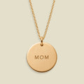 A gold round pendant necklace engraved with the word 'MOM', suspended from a delicate gold chain against a light beige background.