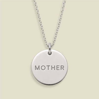 A silver round disc pendant necklace engraved with the word 'MOTHER' in capital letters, suspended from a silver chain against a light beige background.
