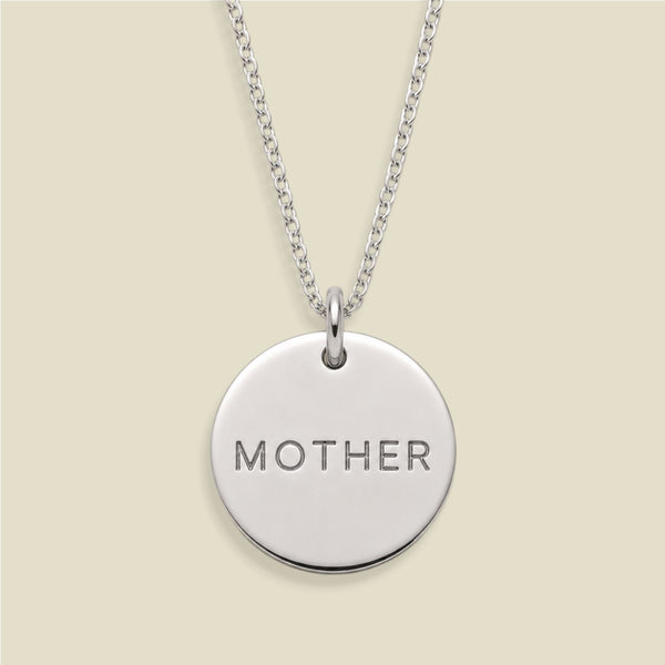 A silver round disc pendant necklace engraved with the word 'MOTHER' in capital letters, suspended from a silver chain against a light beige background.