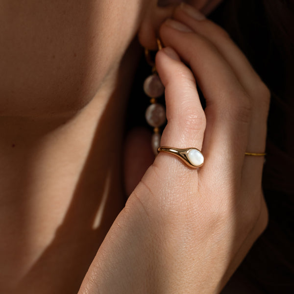 Close-up of a hand wearing a gold ring with an bezel set pearl, gently touching a dangling pearl earring.