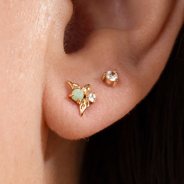 Close-up of a person's ear wearing gold stud earring with a green prong-set stone and an accompanying diamond stud.