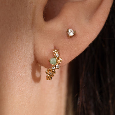 Close-up of a woman's ear with a gold hoop earring with florals and a green prong-set stone and an accompanying diamond stud.
