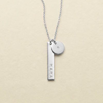 A silver necklace featuring a vertical bar pendant engraved with the word 'MAMA' in uppercase letters and a small round disc charm engraved with the number '5', displayed on a light beige textured background.