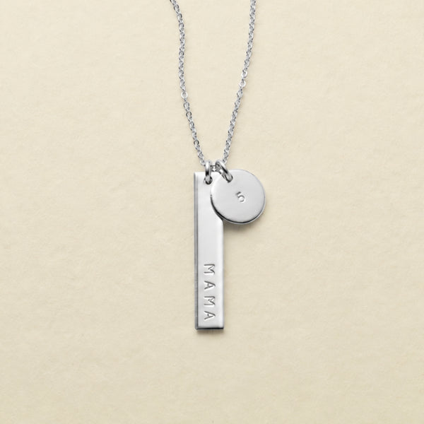 A silver necklace featuring a vertical bar pendant engraved with the word 'MAMA' in uppercase letters and a small round disc charm engraved with the number '5', displayed on a light beige textured background.