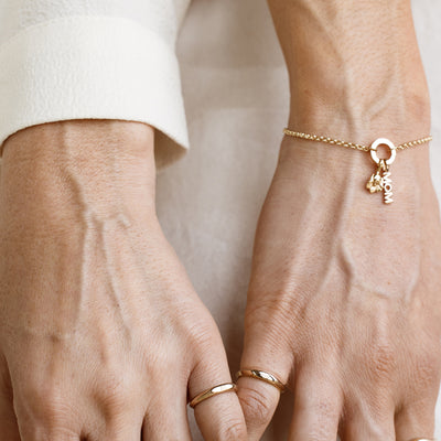 Close-up of hands wearing gold rings and a bracelet with a "MOM" charm on a neutral background