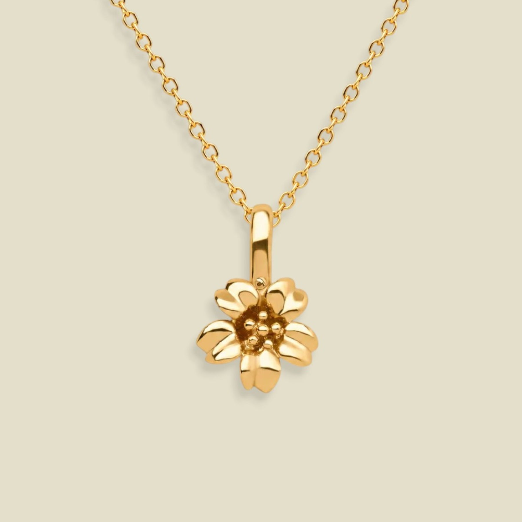 A gold cherry blossom flower pendant necklace on a delicate gold chain, set against a solid light beige background.