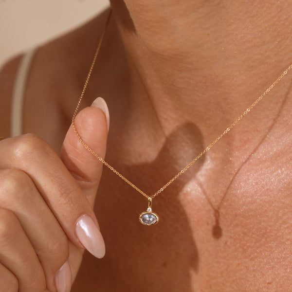 A close-up lifestyle shot of a delicate gold chain necklace with a small oval pendant featuring a light blue-gray stone, worn by a model.