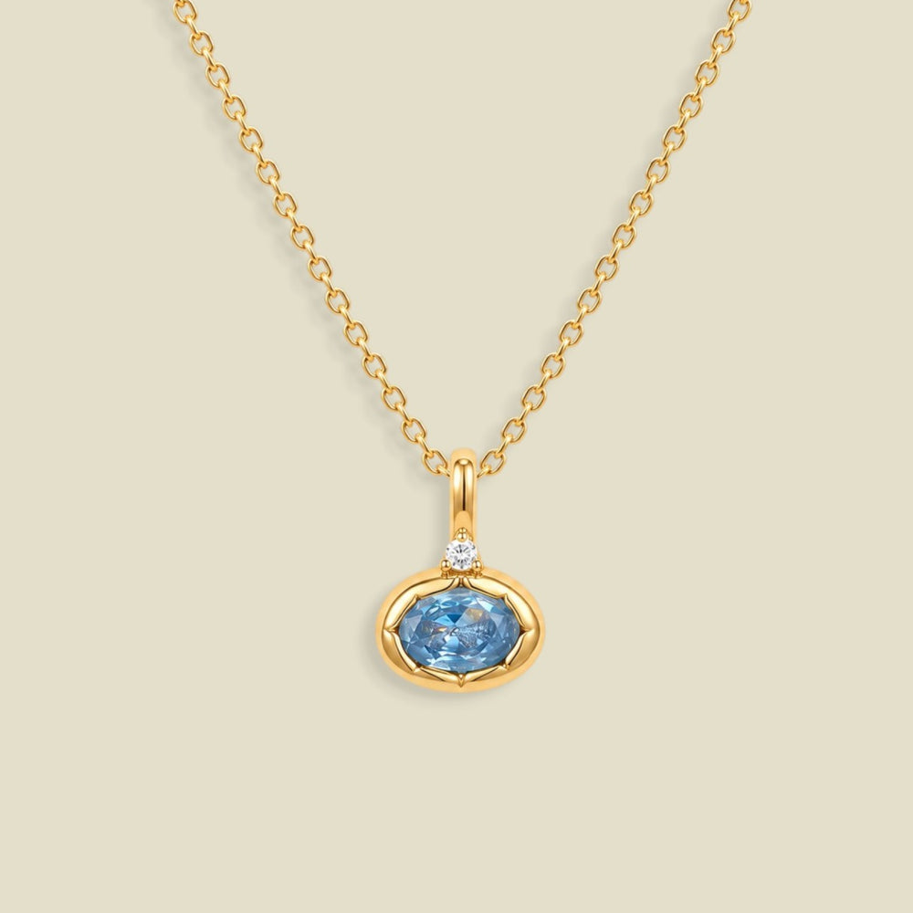 A gold chain necklace with a pendant featuring a small clear stone and an oval bezel set light blue gemstone below it, set against a solid beige background.