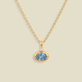 A gold chain necklace with a pendant featuring a small clear stone and an oval bezel set light blue gemstone below it, set against a solid beige background.
