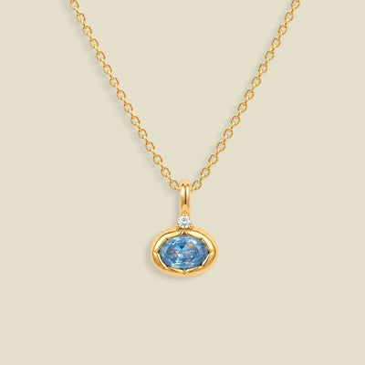 A gold chain necklace with a pendant featuring a small clear stone and an oval bezel set light blue gemstone below it, set against a solid beige background.
