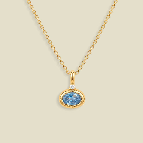 A gold chain necklace with a pendant featuring a small clear stone and an oval bezel set light blue gemstone below it, set against a solid beige background.