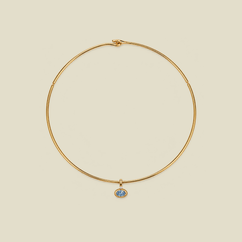 A delicate gold wire circle necklace with a small oval blue stone pendant, displayed on a solid light beige background.