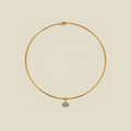 A delicate gold wire circle necklace with a small oval blue stone pendant, displayed on a solid light beige background.