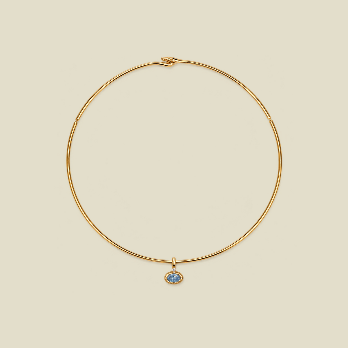 A delicate gold wire circle necklace with a small oval blue stone pendant, displayed on a solid light beige background.
