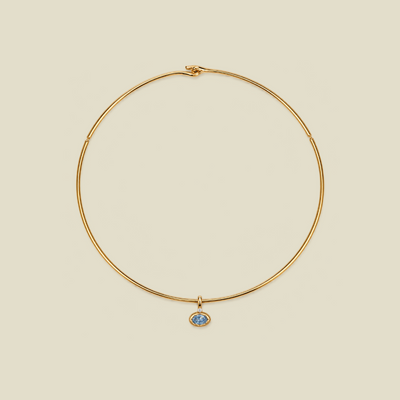 A delicate gold wire circle necklace with a small oval blue stone pendant, displayed on a solid light beige background.
