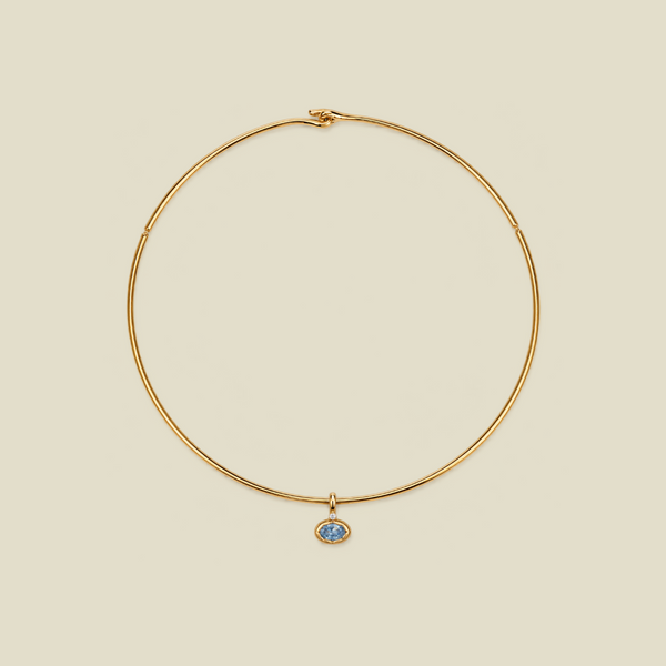 A delicate gold wire circle necklace with a small oval blue stone pendant, displayed on a solid light beige background.
