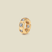 A gold ring charm with multiple bezel set square-cut light grey-blue gemstones set side by side to create the band, shown on a solid beige background.
