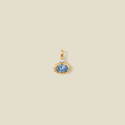 A gold oval pendant charm with a light blue bezel-set oval gemstone and a clear accent stone on top, set against a solid beige background.