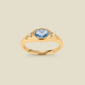 A gold signet ring with a light blue center oval bezel-set gemstone, and two small clear accent stones on either side, set against a solid beige background.