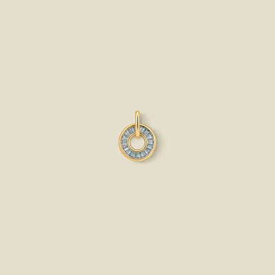 A gold circular pendant featuring light blue baguette-cut stones around a hollow center, displayed against a solid light beige background.