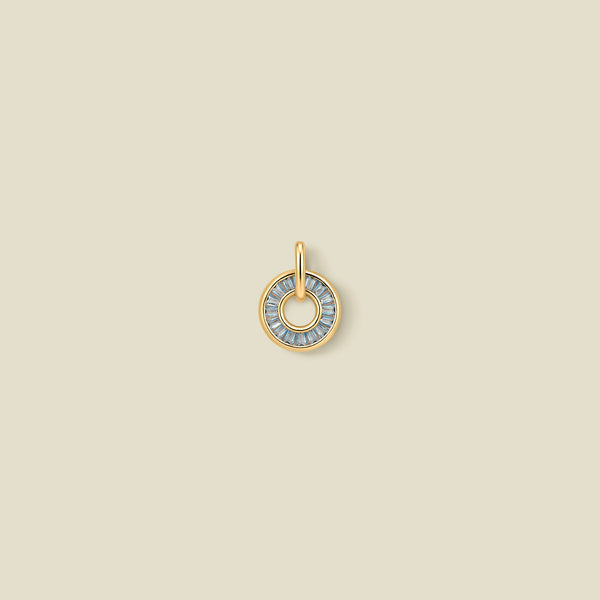 A gold circular pendant featuring light blue baguette-cut stones around a hollow center, displayed against a solid light beige background.