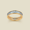 A gold eternity ring featuring a continuous channel setting of baguette-cut light blue gemstones, shown on a solid light beige background.