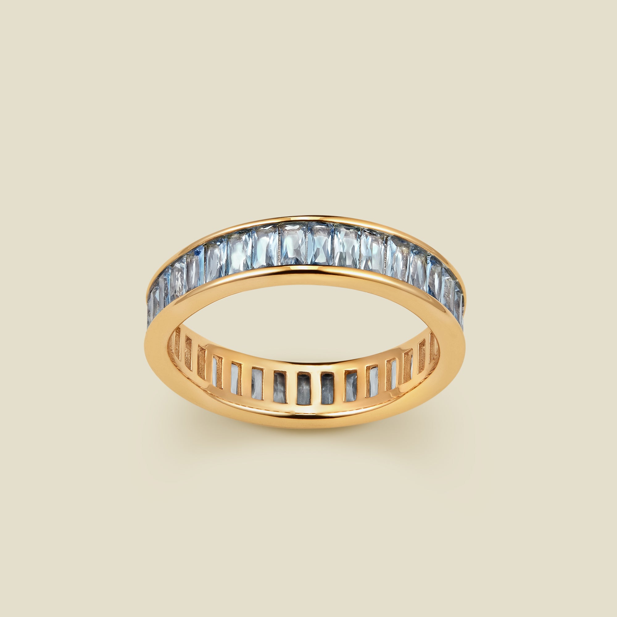 A gold eternity ring featuring a continuous channel setting of baguette-cut light blue gemstones, shown on a solid light beige background.