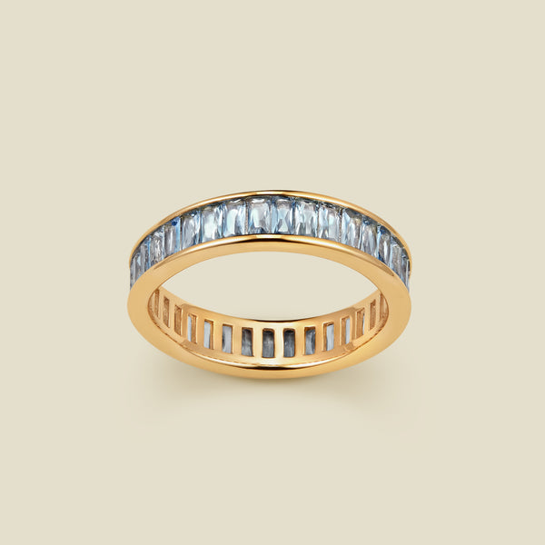 A gold eternity ring featuring a continuous channel setting of baguette-cut light blue gemstones, shown on a solid light beige background.