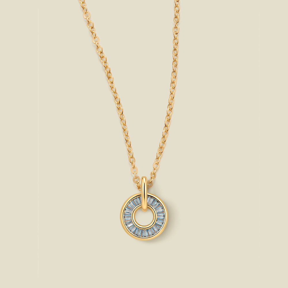 A gold chain necklace with a circular pendant featuring light blue baguette-cut stones around a hollow center, displayed against a solid light beige background.