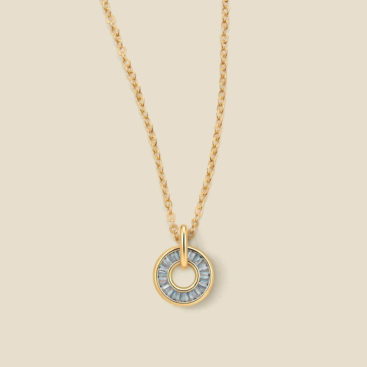 A gold chain necklace with a circular pendant featuring light blue baguette-cut stones around a hollow center, displayed against a solid light beige background.