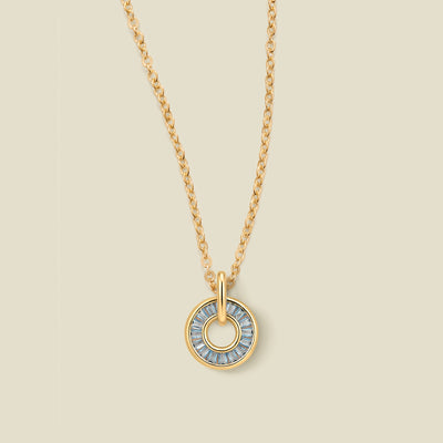 A gold chain necklace with a circular pendant featuring light blue baguette-cut stones around a hollow center, displayed against a solid light beige background.