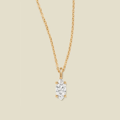 A yellow gold necklace with a single marquise-cut clear stone pendant, displayed against a solid light beige background.