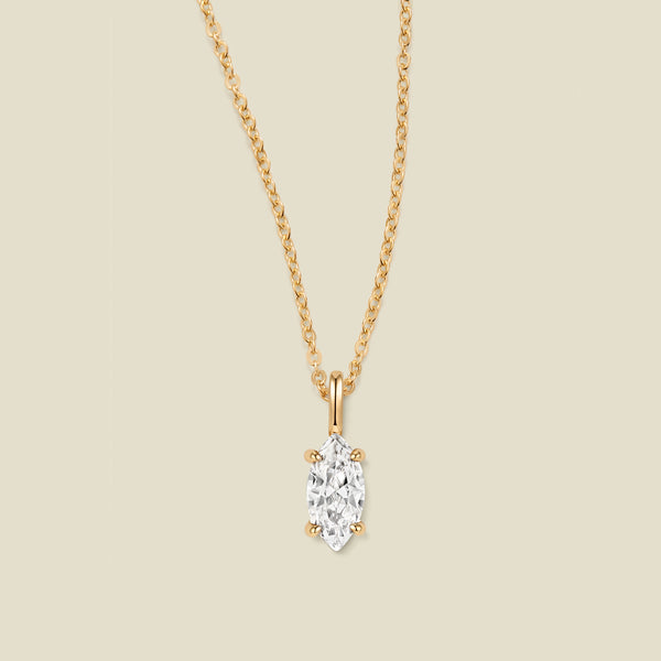 A yellow gold necklace with a single marquise-cut clear stone pendant, displayed against a solid light beige background.