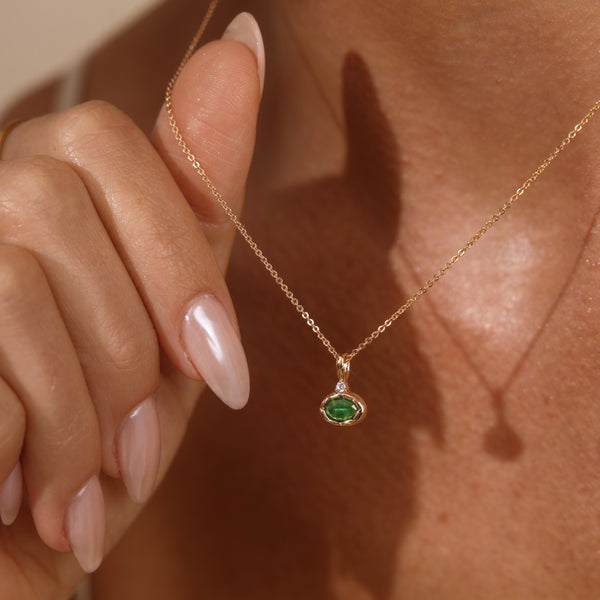 A close-up lifestyle shot of a person wearing a delicate gold chain necklace with a small oval green gemstone pendant, held gently by a hand with manicured nails.