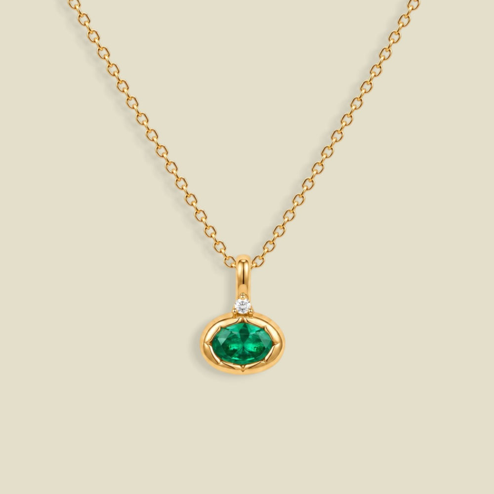 A gold chain necklace with a small oval pendant featuring a dark emerald green faceted gemstone and a tiny clear accent stone above it, shown on a light beige background.