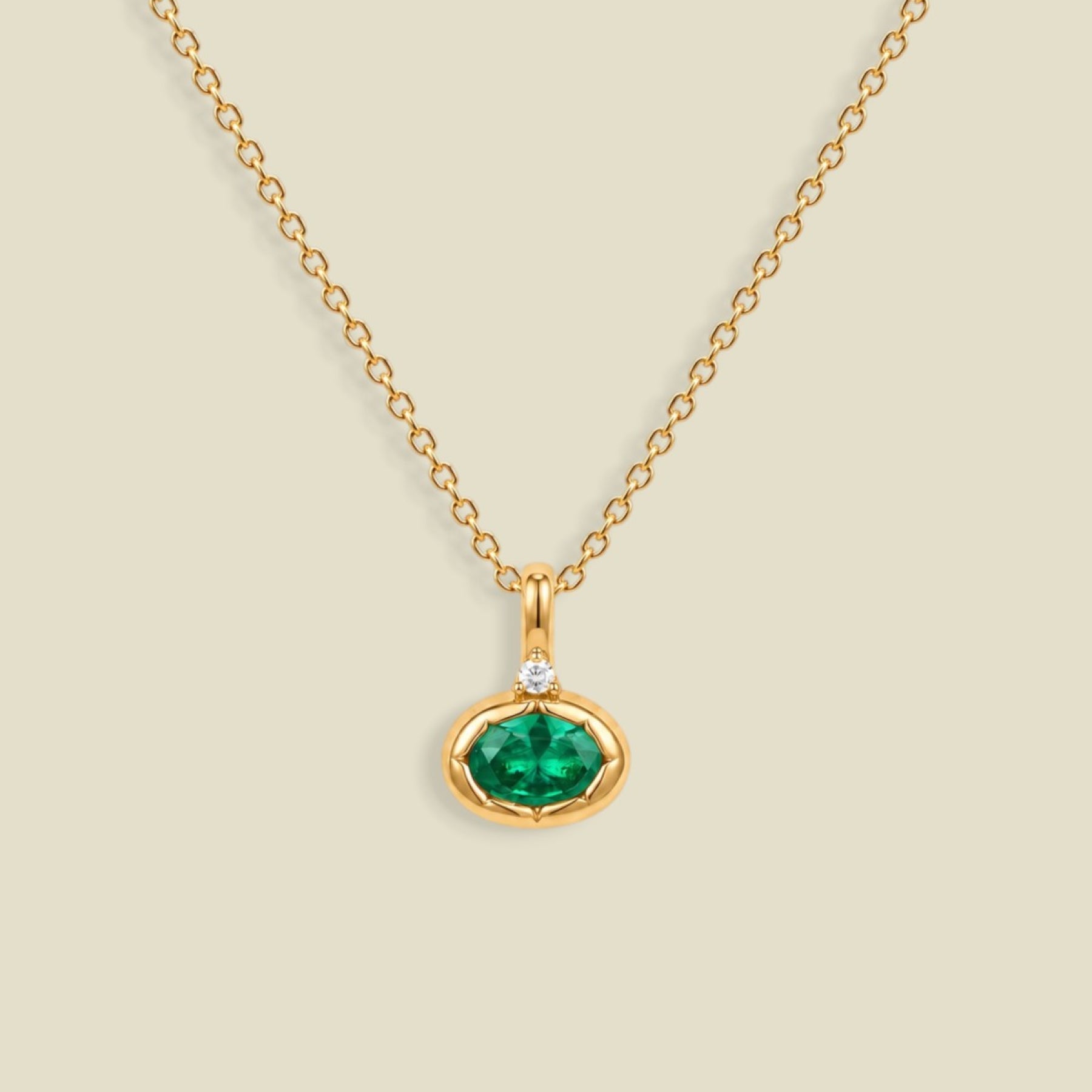 A gold chain necklace with a small oval pendant featuring a dark emerald green faceted gemstone and a tiny clear accent stone above it, shown on a light beige background.