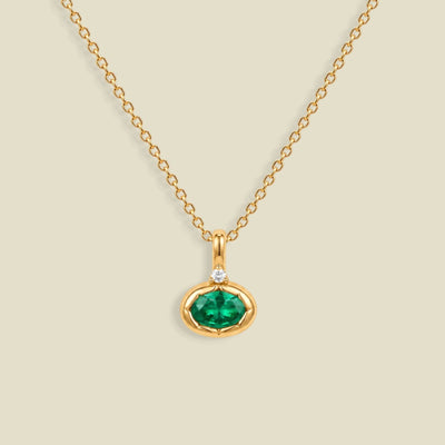 A gold chain necklace with a small oval pendant featuring a dark emerald green faceted gemstone and a tiny clear accent stone above it, shown on a light beige background.