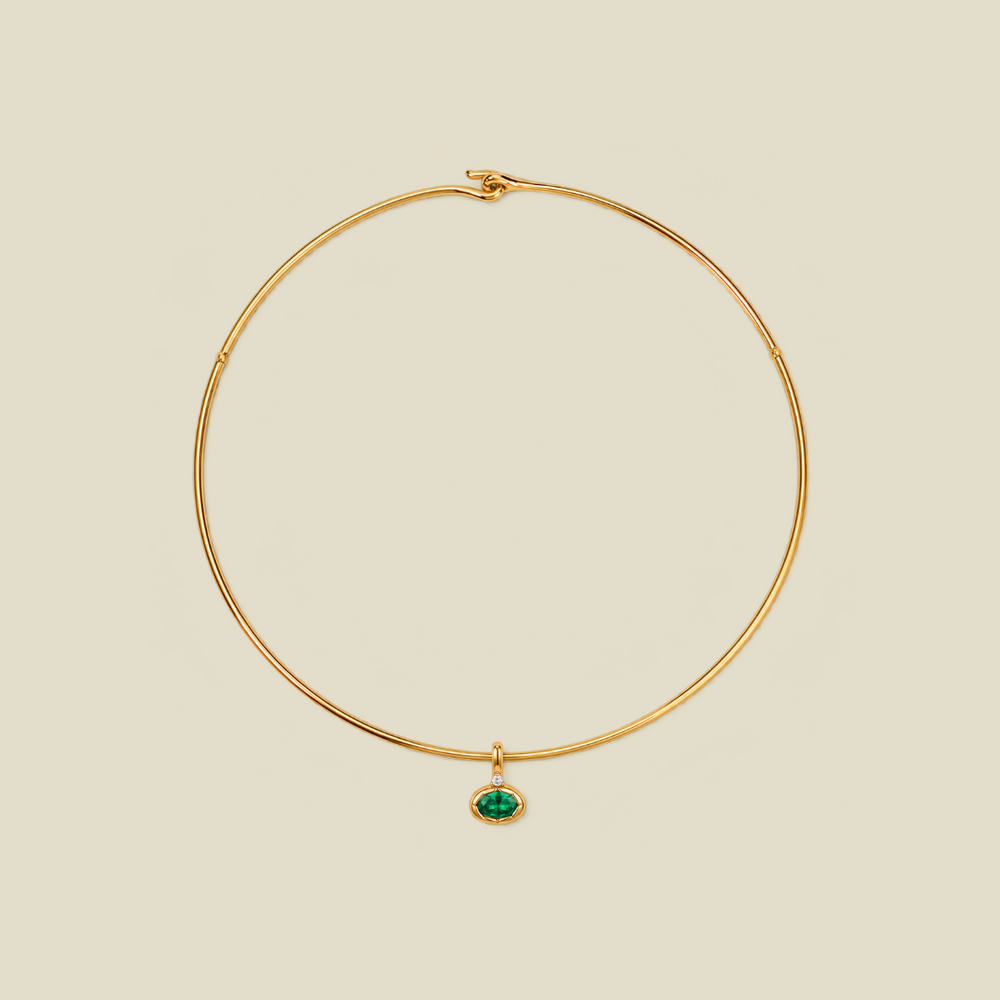 A delicate gold wire necklace with a small oval green gemstone pendant, displayed on a solid light beige background.
