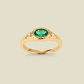 A gold signet ring with a green center oval bezel-set gemstone, and two small clear accent stones on either side, set against a solid beige background.