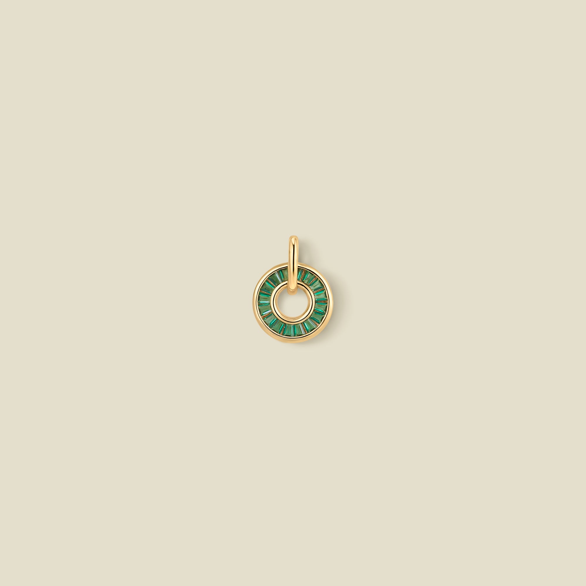 A gold circular pendant featuring dark green baguette-cut stones around a hollow center, displayed against a solid light beige background.