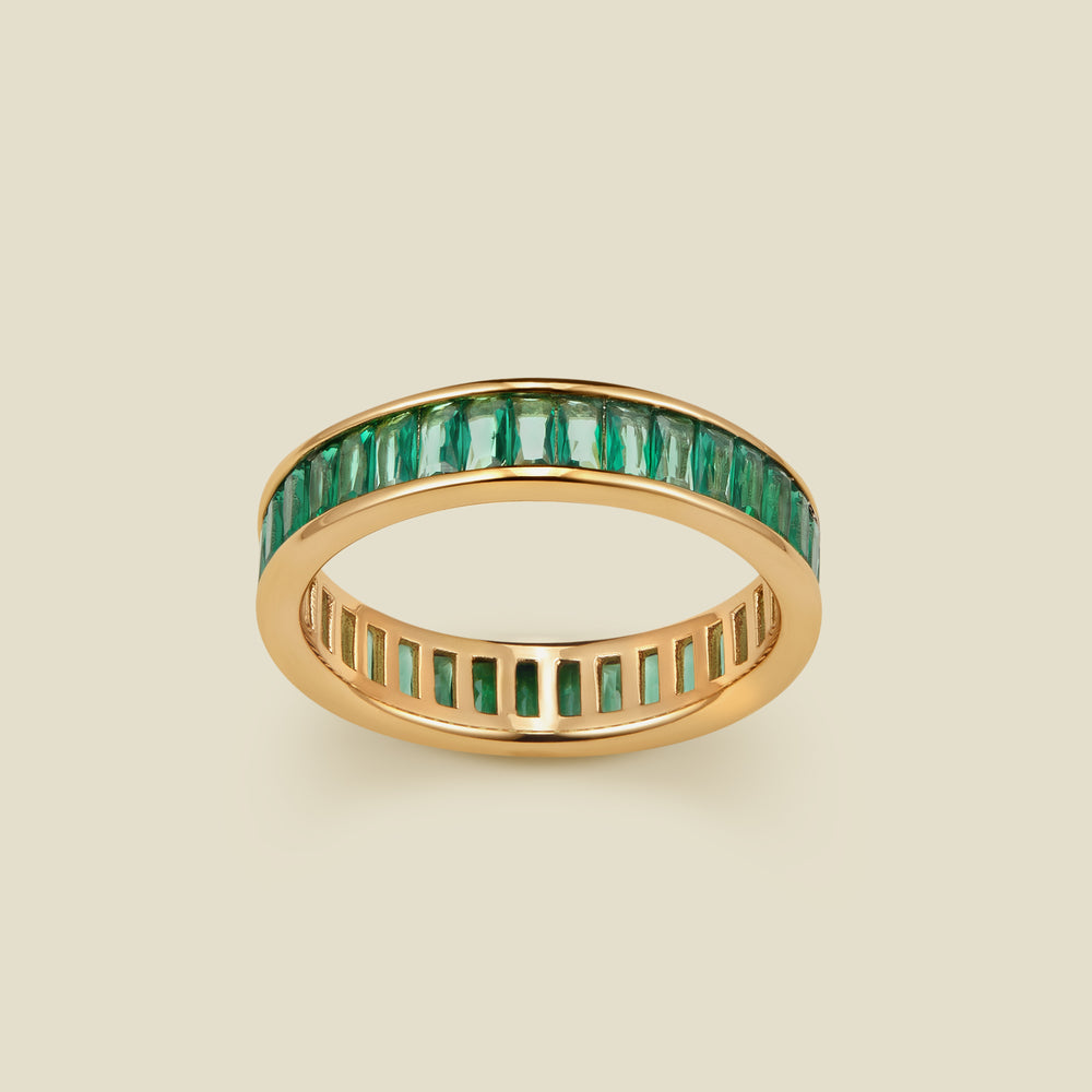 A gold eternity ring featuring a continuous channel setting of baguette-cut dark green gemstones, shown on a solid light beige background.