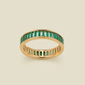 A gold eternity ring featuring a continuous channel setting of baguette-cut dark green gemstones, shown on a solid light beige background.