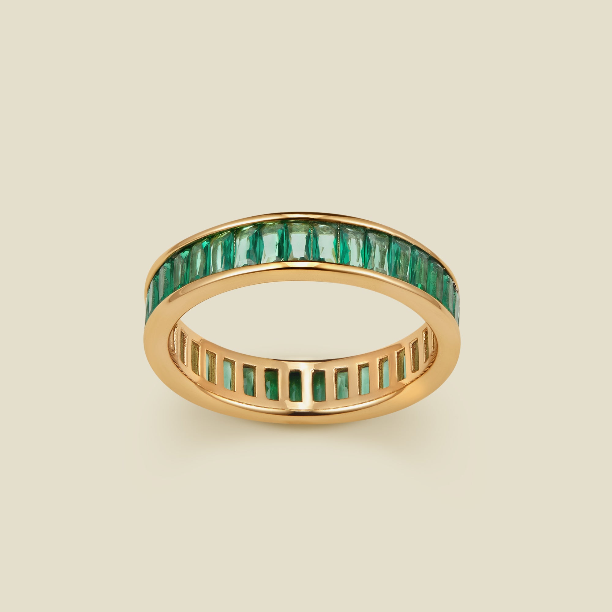 A gold eternity ring featuring a continuous channel setting of baguette-cut dark green gemstones, shown on a solid light beige background.