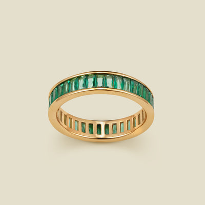 A gold eternity ring featuring a continuous channel setting of baguette-cut dark green gemstones, shown on a solid light beige background.