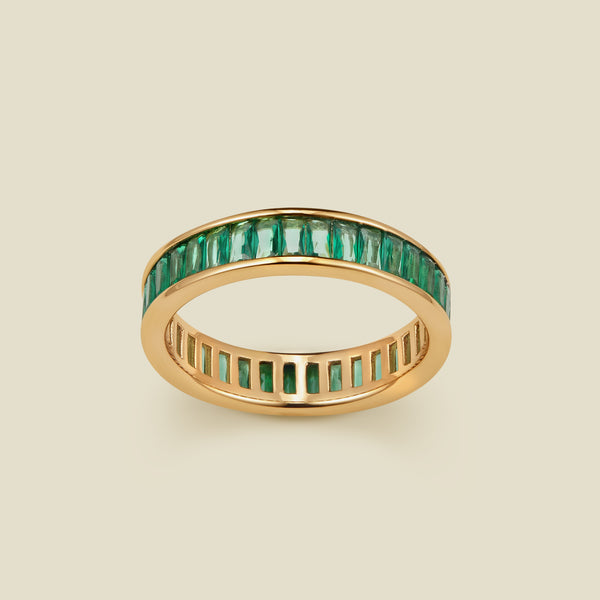 A gold eternity ring featuring a continuous channel setting of baguette-cut dark green gemstones, shown on a solid light beige background.