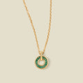 A gold chain necklace with a circular pendant featuring dark green baguette-cut stones around a hollow center, displayed against a solid light beige background.