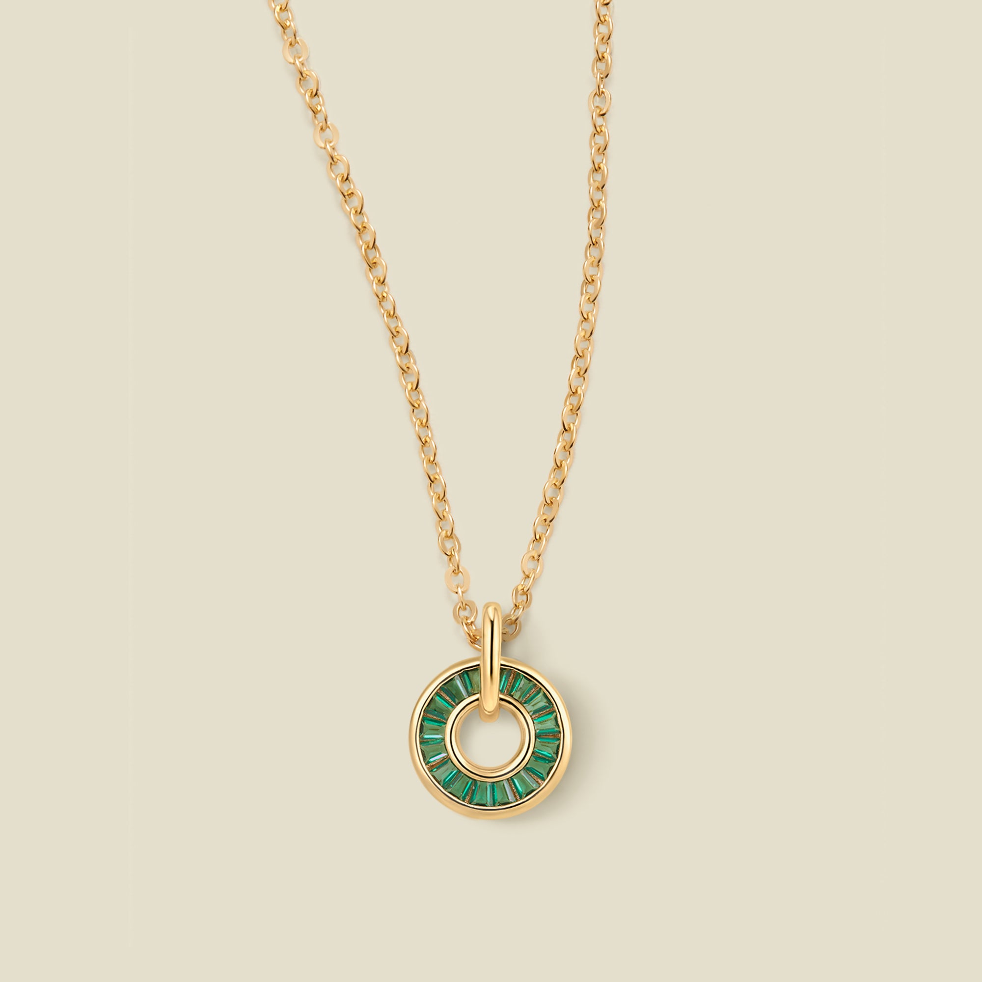 A gold chain necklace with a circular pendant featuring dark green baguette-cut stones around a hollow center, displayed against a solid light beige background.